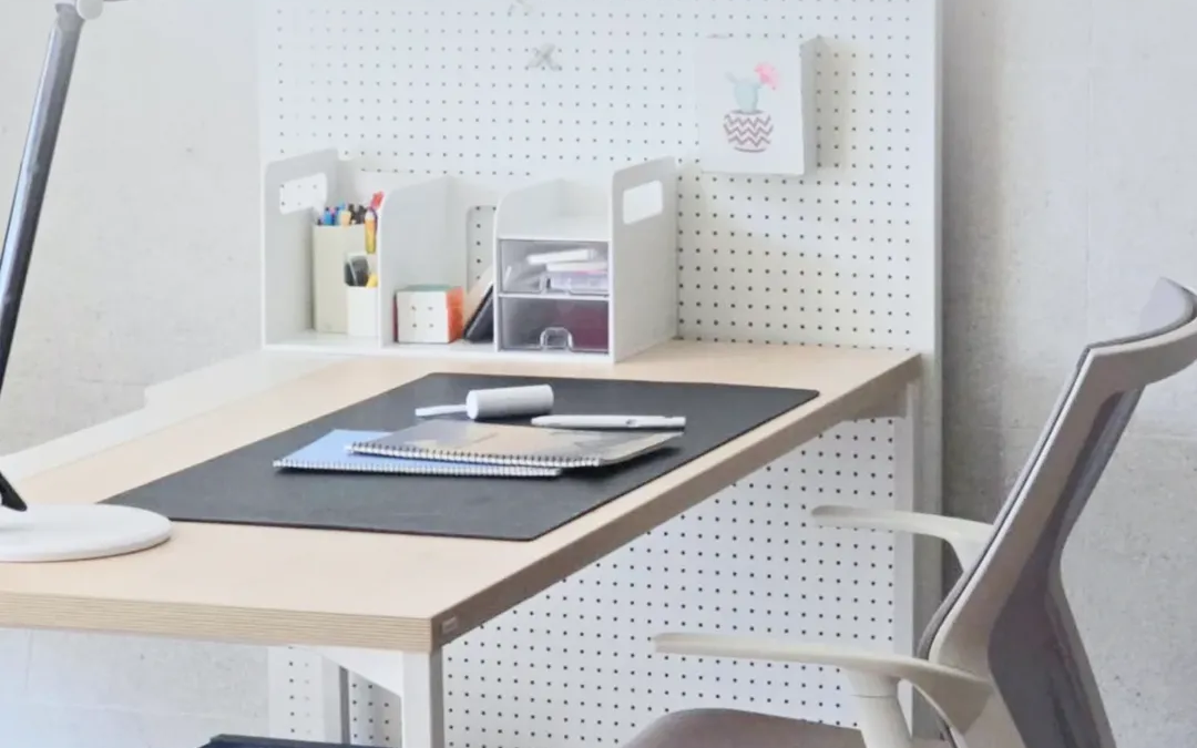 Meja kerja putih minimalis dengan papan pegboard, lampu meja, dan buku catatan