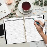 Planner spiral tahun 2027 di meja putih dengan tangan memegang pena warna teal