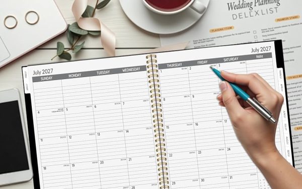 Planner spiral tahun 2027 di meja putih dengan tangan memegang pena warna teal