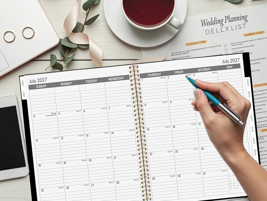 Planner spiral tahun 2027 di meja putih dengan tangan memegang pena warna teal