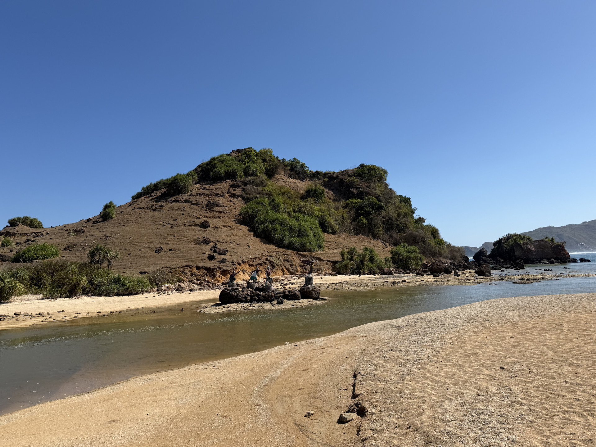 Pantai tropis berpasir dengan air tenang dan bukit hijau di kejauhan