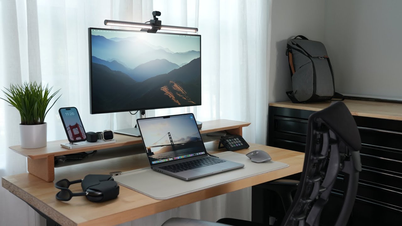 Meja kerja kayu rapi dengan monitor, laptop, telepon, headphone, dan tanaman
