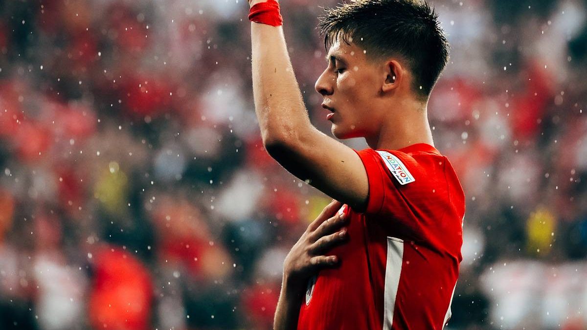 Pemain muda berjersey merah merayakan gol dengan tangan menunjuk ke atas di stadion