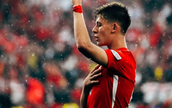 Pemain muda berjersey merah merayakan gol dengan tangan menunjuk ke atas di stadion
